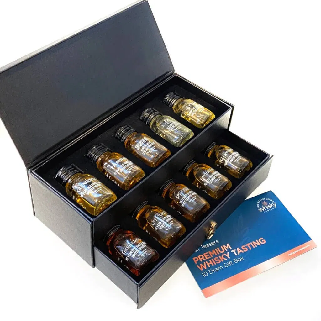 A luxury black gift box is opened to reveal ten 30ml miniature bottles, or drams, of premium whisky arranged neatly in a dark foam insert. The bottles are labeled with different distillery names, suggesting a variety of scotch or international whiskies. A colourful guide card titled '10 Dram Gift Box' is visible to the side. Ideal whisky sampling gift for birthdays, Father's Day, or corporate gifting.