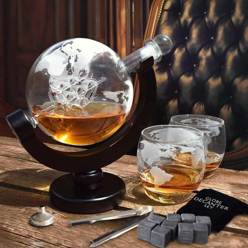 A premium globe-shaped glass decanter on a curved dark wood stand is half-filled with amber spirit, showcasing an intricate glass sailing ship model inside. The decanter is accompanied by two matching globe-etched whisky glasses and bar accessories, including chilling stones and tongs. The set is displayed on a rustic wooden surface in a luxurious, leather-backed armchair setting, suggesting travel, adventure, and sophistication. Ideal gift for a traveller, whisky enthusiast, or man cave.