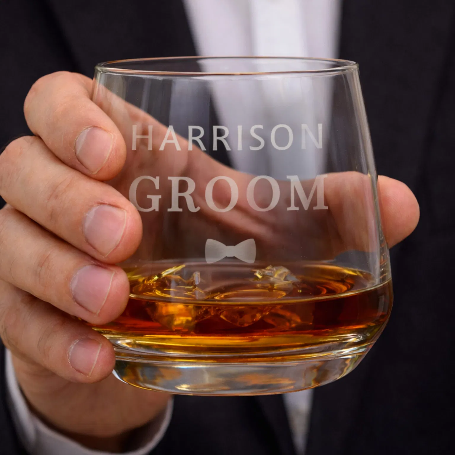 Personalised glass tumbler held by a man in a suit, engraved with 'Harrison Groom' and a bowtie icon, filled with whisky and ice.