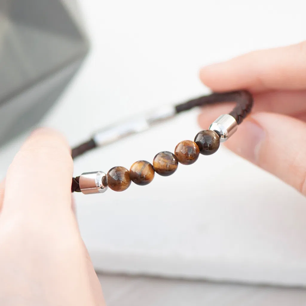 A close-up of a men's bracelet featuring a dark brown woven leather band, a central section of polished tiger's eye gemstone beads, and a curved silver-toned metal clasp engraved with the text "GADGET MAN RD," held between two hands against a blurred background.