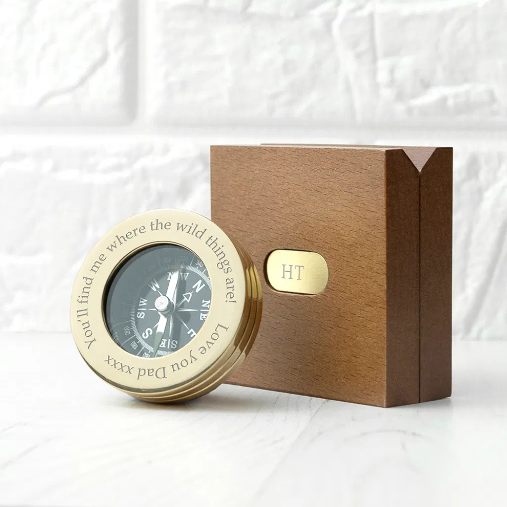A polished brass compass engraved with the message 'You’ll find me where the wild things are! Love you Dad xxxx' resting next to a monogrammed wooden gift box with 'HT' engraved on a brass plate.