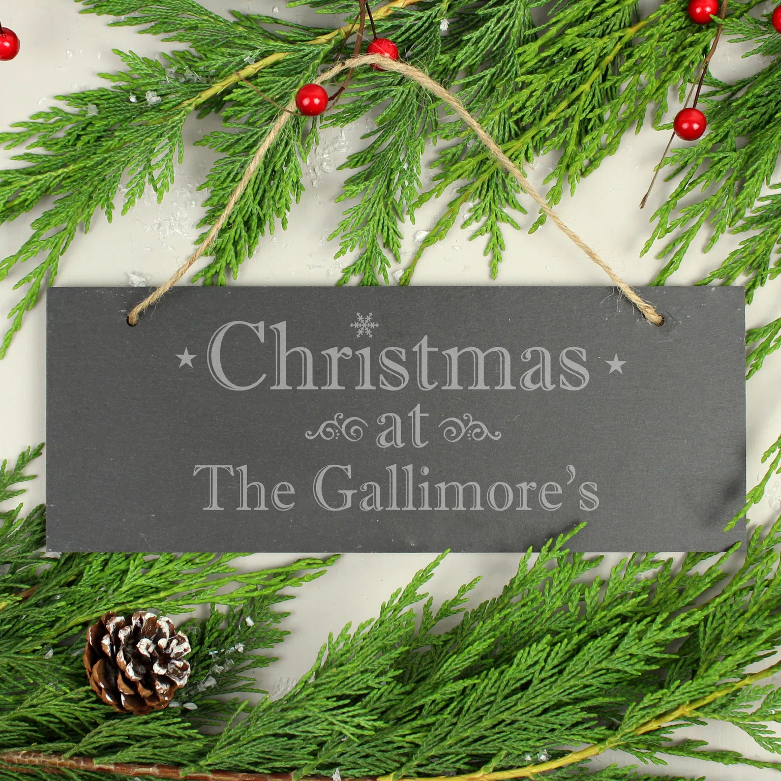 A rectangular dark natural slate sign is centered flat against a light background, hanging from a piece of rustic jute twine. The sign is laser-engraved with stars, a snowflake, and the fixed text 'Christmas at' above the custom name 'The Gallimore's'. It is bordered along the top and bottom by fresh green pine branches with red winter berries and a pinecone.