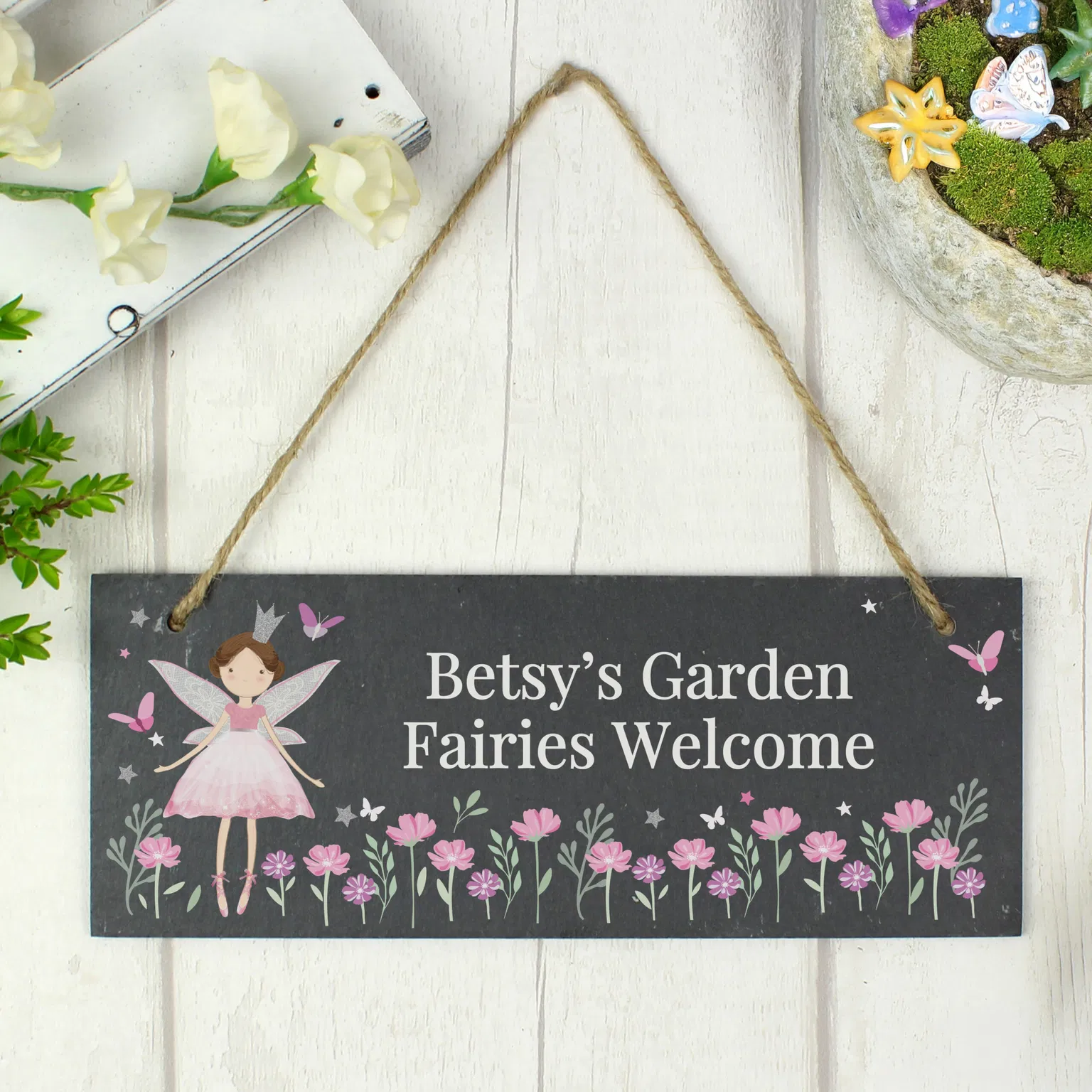 A rectangular natural dark slate hanging sign hangs from a rustic jute twine loop against a white wood panel background. The left features a printed fairy princess illustration next to the custom text 'Betsy's Garden, Fairies Welcome' above a border of pink wildflowers and butterflies. A miniature fairy garden container is partially visible in the top right corner.