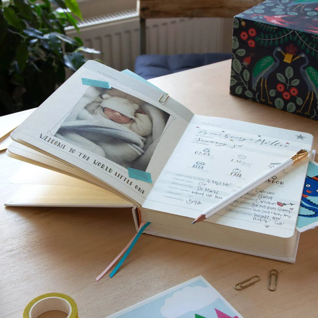 A beautifully designed, open baby journal or memory book, displaying the first page with a photo of a newborn baby wrapped in a blanket. The page includes fill-in sections for the baby's name ('George Miller'), birth details (date, weight, eye colour), and early milestones. Ideal keepsake gift for new parents to record their child's first 26 months.
