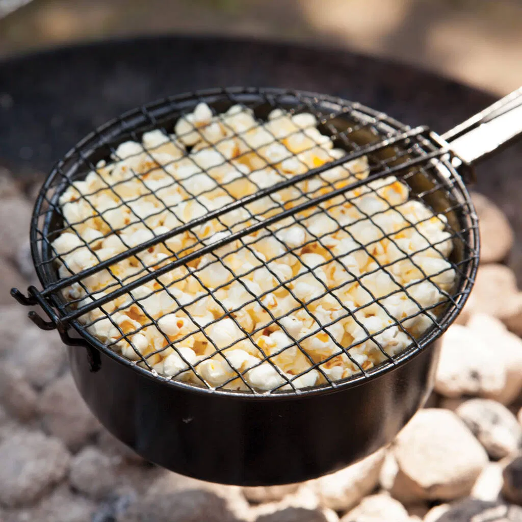 A round, black metal popcorn pan with a long handle and a secure mesh lid, holding freshly popped white popcorn kernels. The pan is held directly over glowing charcoal briquettes in an outdoor barbecue or fire pit. The close-up view emphasizes the rugged cooking tool and the successful, fluffy result. Ideal accessory for camping, grilling, or outdoor entertaining.