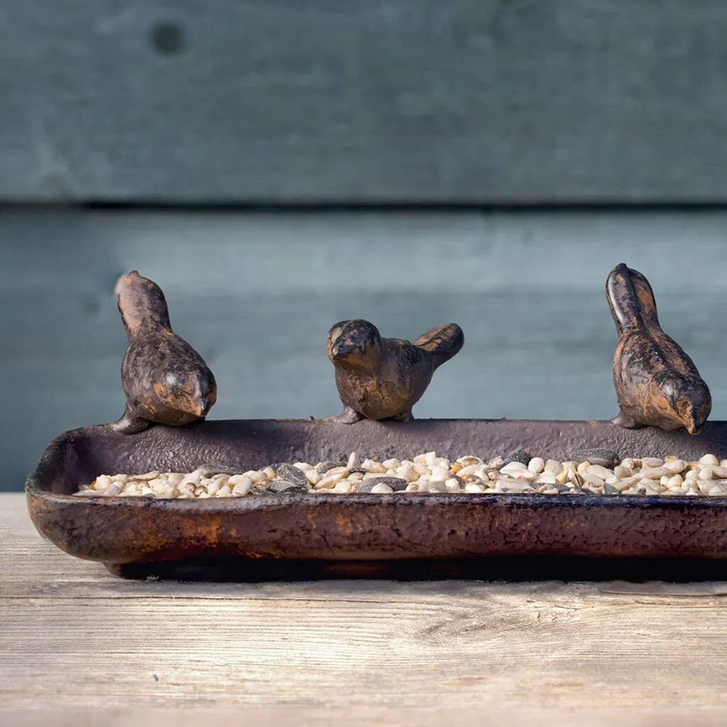 Close-up of a rustic, cast iron bird feeder tray filled with sunflower seeds and nuts, featuring three decorative bird sculptures perched on the rim. The feeder sits on a wooden surface against a blurred blue wooden fence background. Ideal wildlife garden gift.