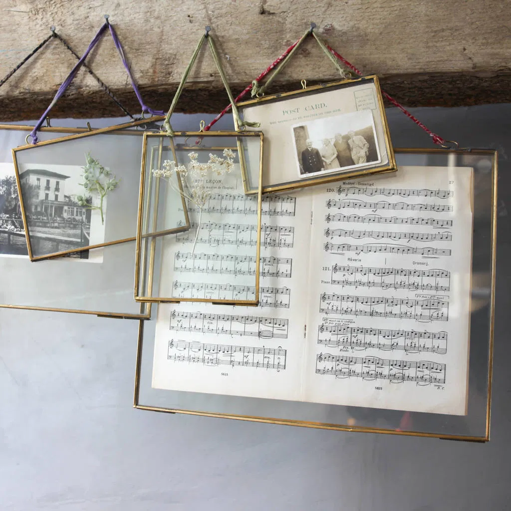 A group of several Brass Hanging Picture Frames of different sizes and orientations (portrait and landscape) suspended from a wooden beam by colourful recycled sari ties. The transparent glass frames display various items, including a vintage postcard, dried white flowers, a botanical print, and antique sheet music, creating a unique, overlapping collage.