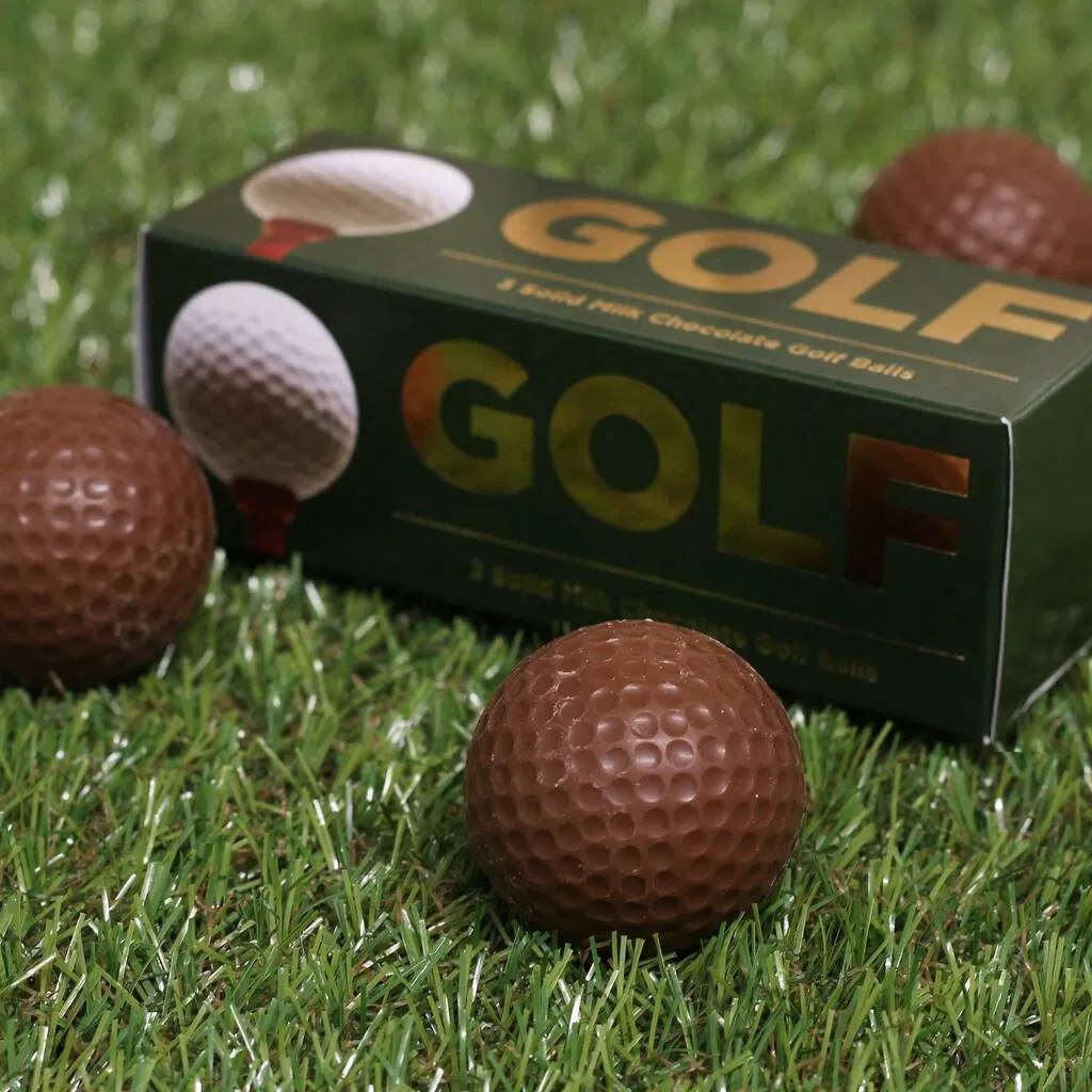 Close-up of solid milk chocolate golf balls sitting on artificial grass with a green gift box.