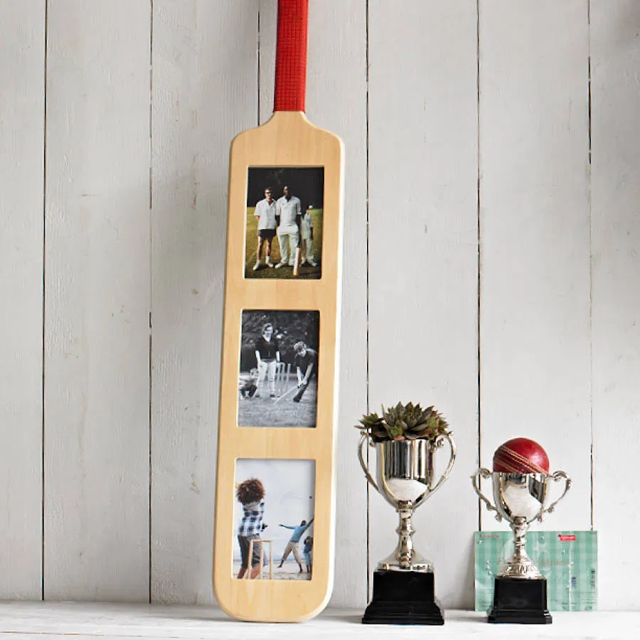 Decorative wooden cricket bat photo frame with a red handle wrap, featuring three vertical rectangular photo slots. Displayed next to silver cricket trophies, emphasizing its use as a unique gift for cricket fans.