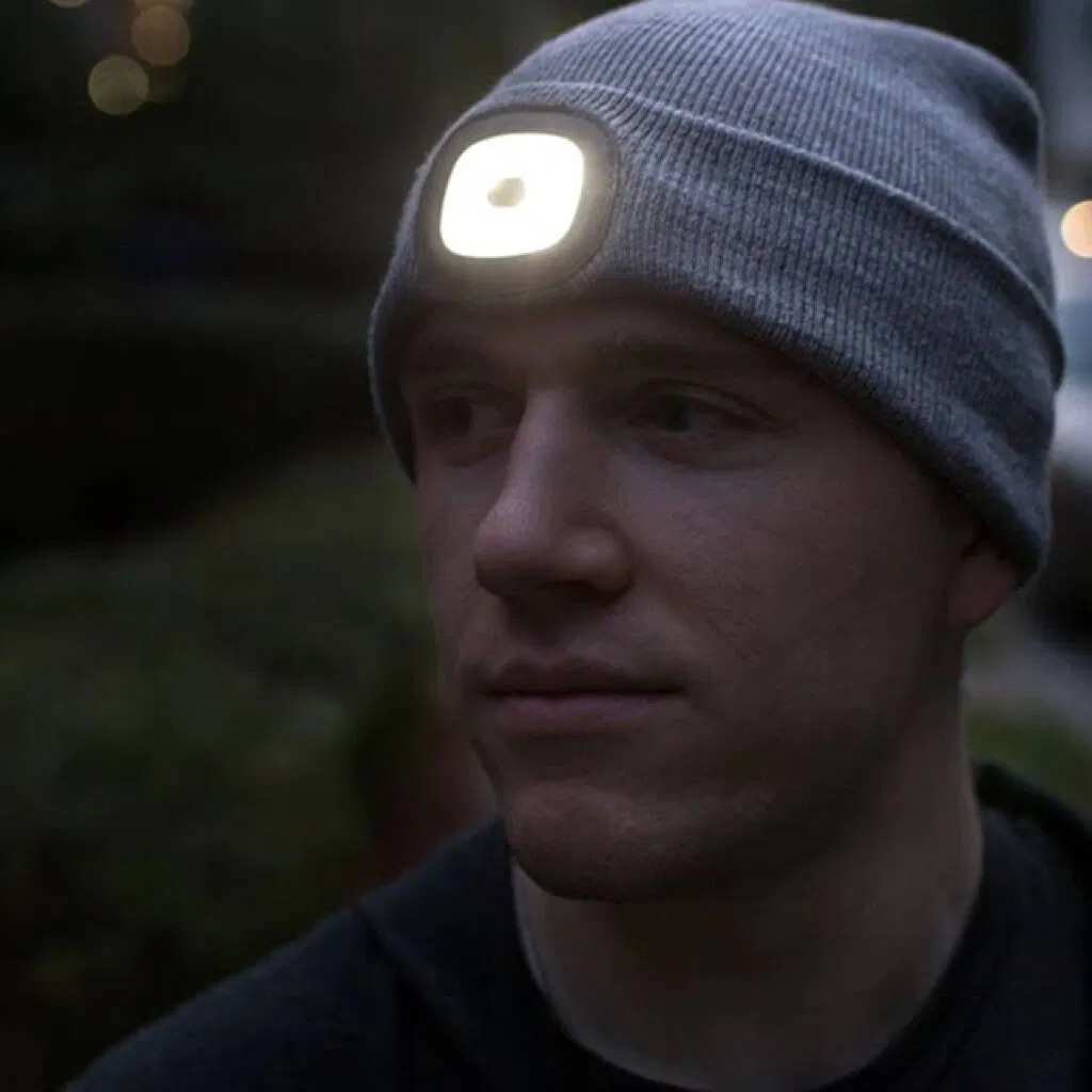 A man wearing a grey knitted beanie hat with a bright, square LED light panel embedded in the forehead, facing forward in a dimly lit, outdoor setting. The light is switched on for visibility. Ideal practical gift for cyclists, runners, hikers, or anyone needing hands-free light in winter.