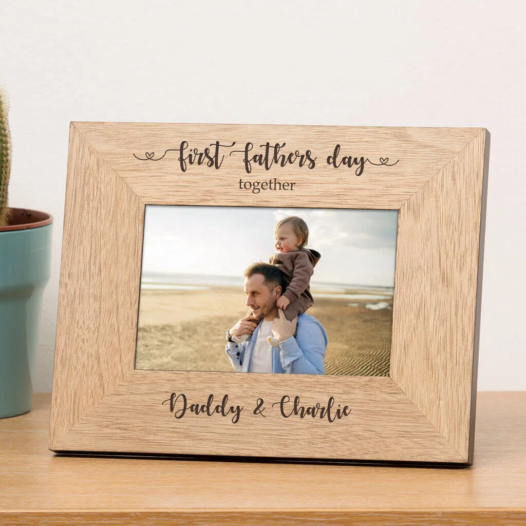 Personalized wooden picture frame engraved with the text 'first fathers day together' at the top and custom names, 'Daddy & Charlie,' at the bottom. The frame holds a photo of a father carrying his baby on his shoulders on a beach.