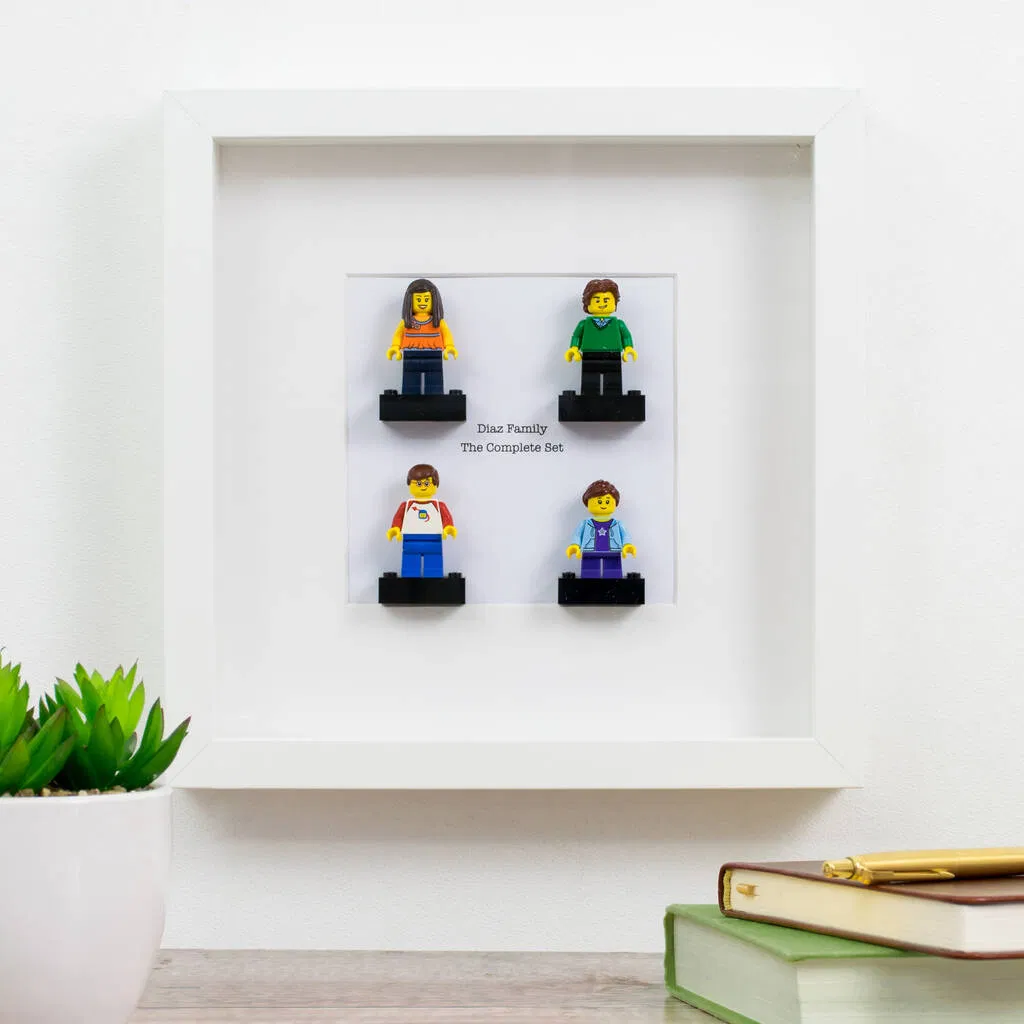 A white-framed display piece featuring four custom mini figures standing on black platforms. Below the figures, the text "Diaz Family The Complete Set" is printed, suggesting a family or group portrait made from personalized toy bricks. The frame is shown sitting on a desk next to a green potted plant and books. Ideal personalised family gift, Mother's Day, Father's Day, or anniversary present for fans of brick toys.