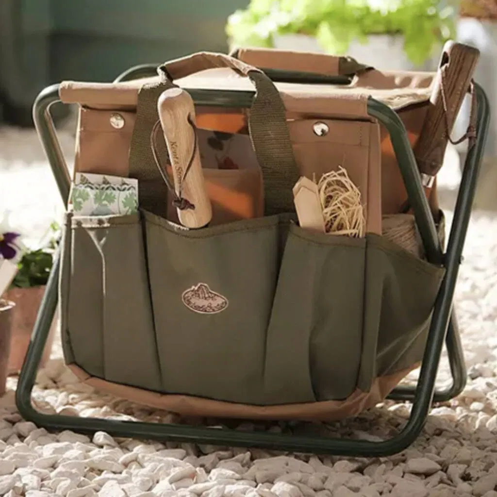 Foldable garden tool stool with a khaki green and brown canvas storage bag attached, displayed on a gravel patio surrounded by terracotta pots, pansies, and colourful flowers. Ideal portable gardening gift and accessory.