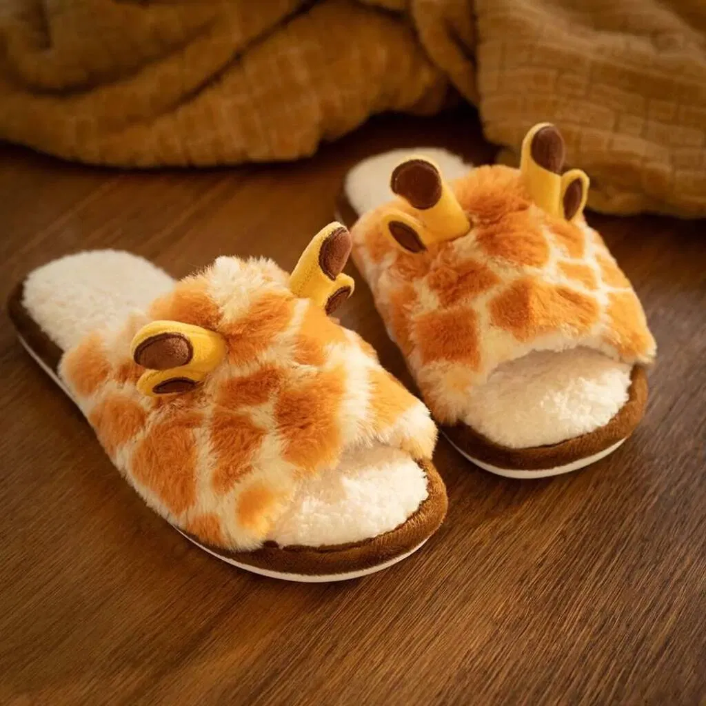 A pair of fluffy, plush giraffe slippers with horns and brown spots, resting on a wooden floor next to a warm, folded blanket.