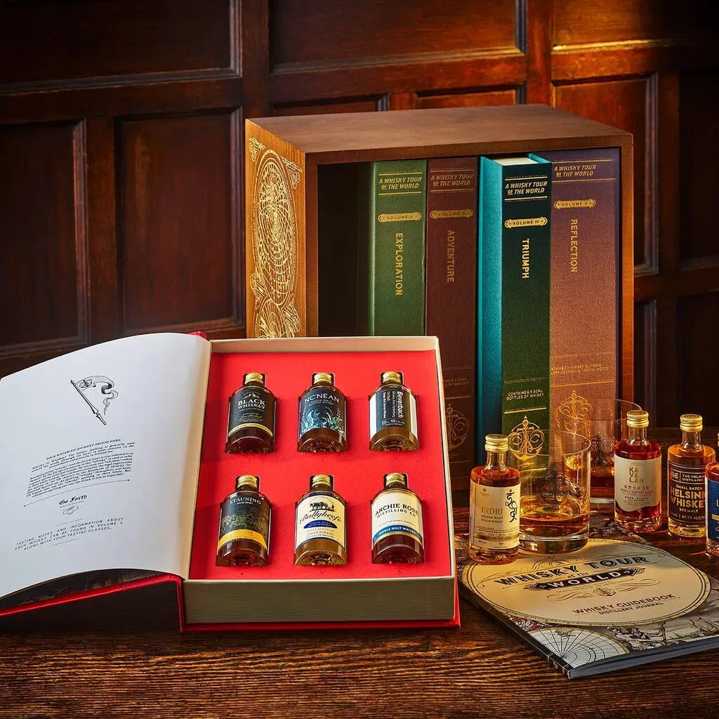 A luxury whisky tasting set featuring eight miniature bottles of diverse whiskies housed in a collector's box designed to look like a set of antique books titled 'Exploration,' 'Adventure,' 'Triumph,' and 'Reflection.' The box is open to display the selection, with a guide book and a Glencairn-style tasting glass nearby, set against a rich wooden background.