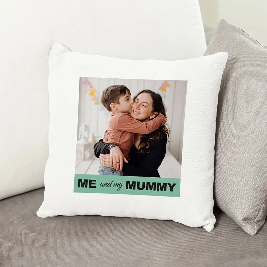 A square white cushion cover featuring a large printed photo of a happy woman (Mum) being hugged and kissed on the cheek by a young boy. Beneath the photo is a green banner with the text "ME and my MUMMY". The cushion is displayed on a grey sofa.