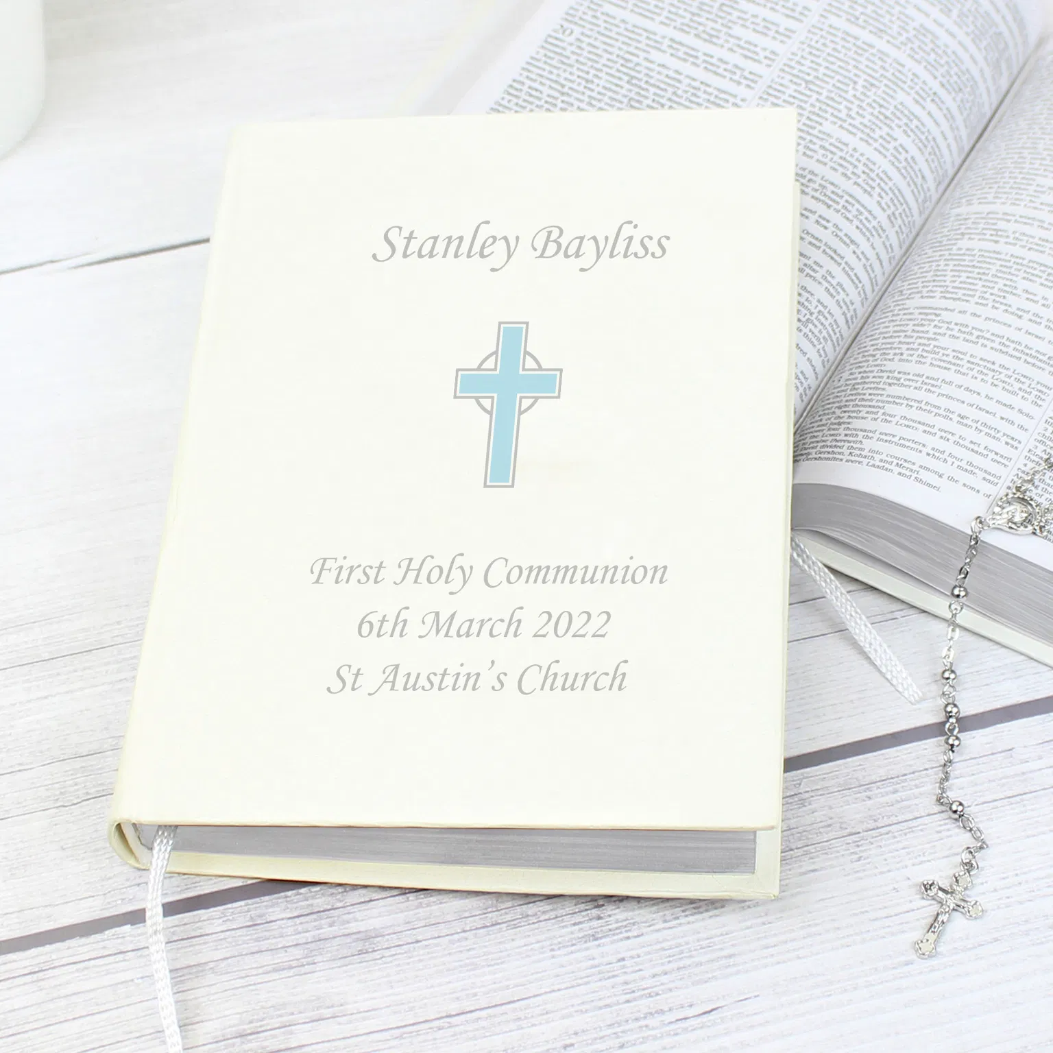 The front cover of the Personalised Eco Friendly Blue Cross Holy Bible from Abode Home & Gifts customised for Stanley Bayliss's First Holy Communion, resting at an angle on a white wood surface next to an open Bible and a silver rosary bead necklace.