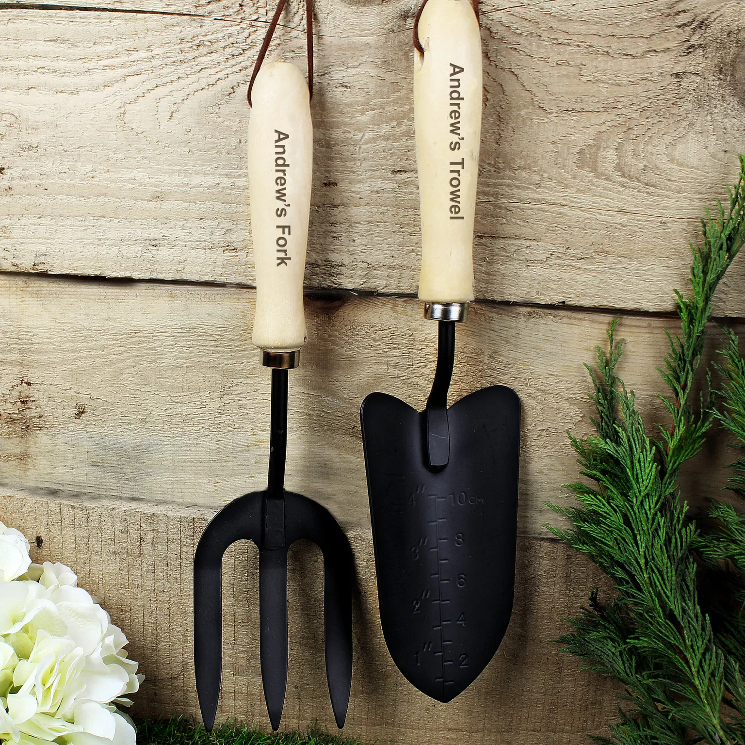 Personalised gardening fork and trowel set with 'Andrew's Fork' and 'Andrew's Trowel' engraved on the wooden handles against a rustic wood backdrop.