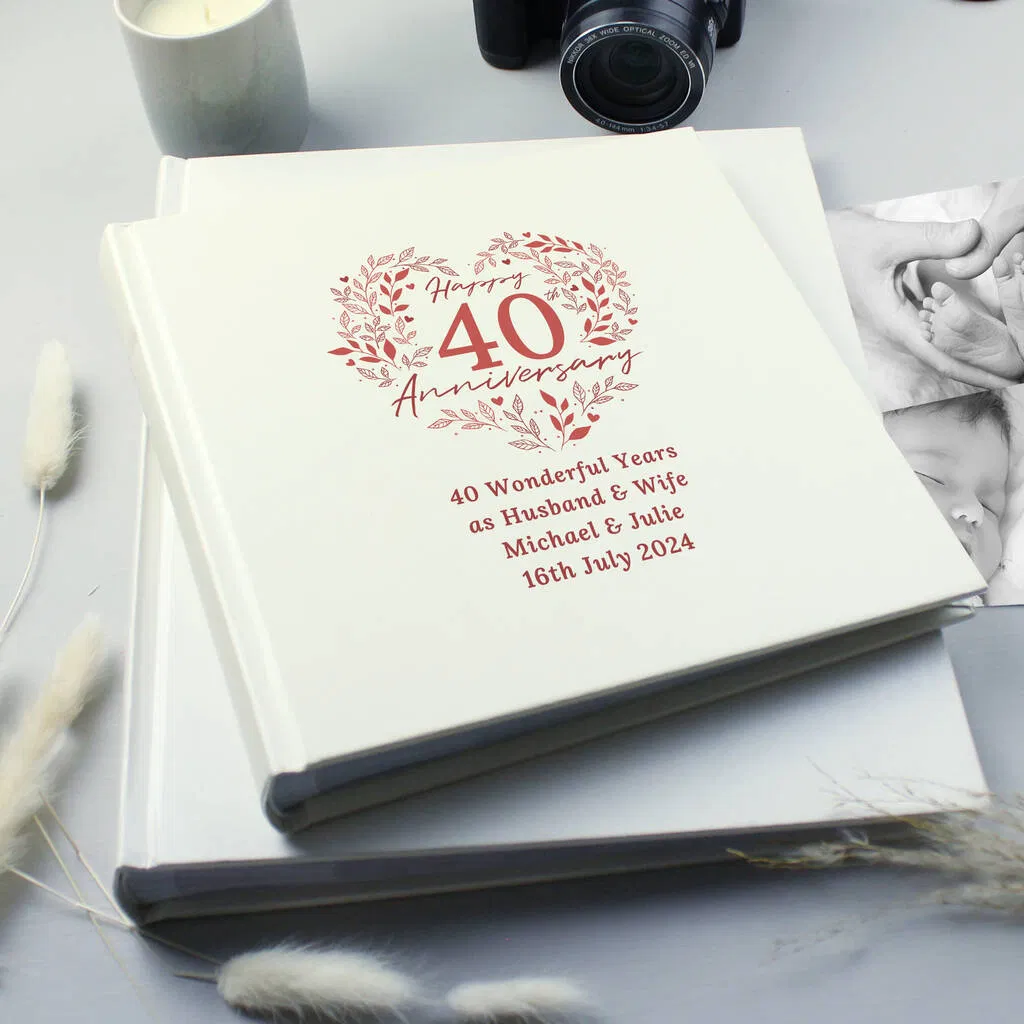 A stack of cream photo albums featuring a personalized design for a 'Happy 40th Anniversary' in ruby red print, including the names of the couple and the date '16th July 2024'. Set on a light grey background with a candle and camera nearby. Ideal personalised Ruby Wedding Anniversary gift.