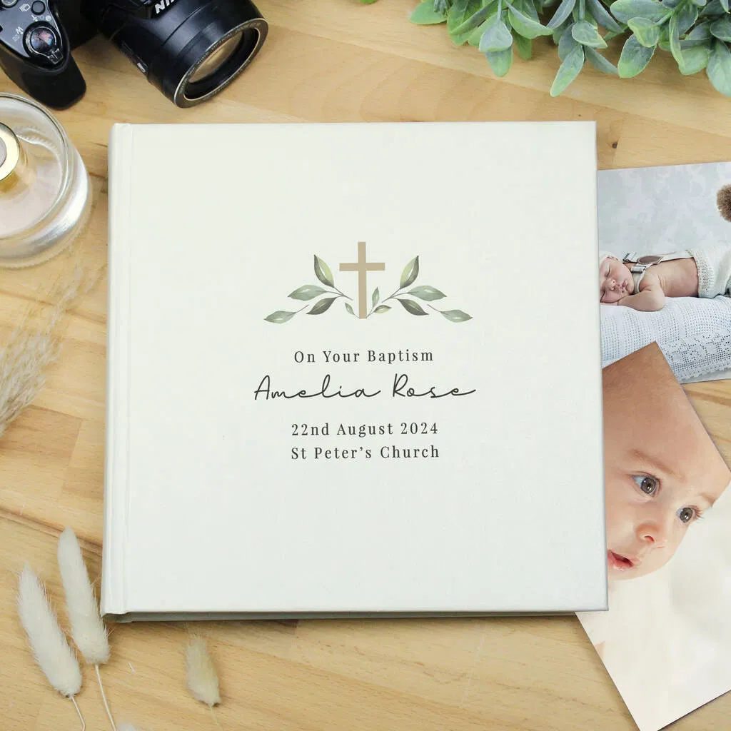 A personalised, cream-coloured photo album for a Baptism, displayed on a light wooden surface. The cover features a subtle design of a cross framed by green leafy sprigs above the text: "On Your Baptism," the name "Amelia Rose" in script, the date "22nd August 2024," and "St Peter's Church." The album is surrounded by a camera lens, a glass bottle, dried reeds, and a couple of small baby photos tucked beneath the cover.