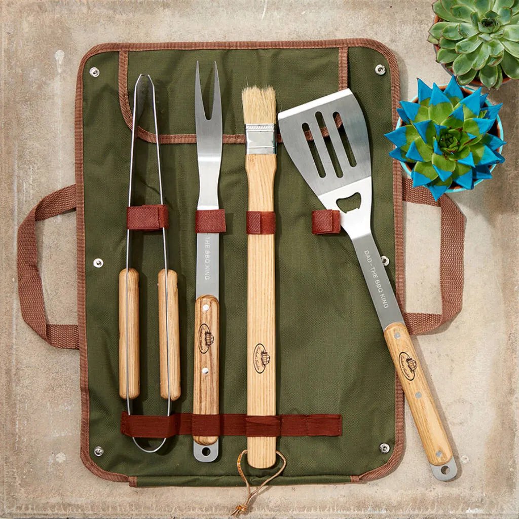 An overhead shot of a personalised barbecue tool set with wooden handles, laid out in an olive green canvas storage roll-up bag. The set includes tongs, a carving fork, a basting brush, and a spatula, all engraved with messages like 'DAD - THE BBQ KING'. Ideal personalised gift for Father's Day or a grill master.