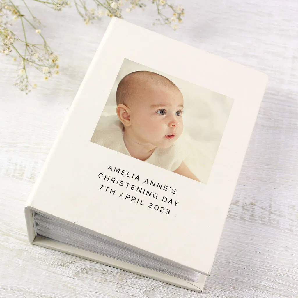 A cream-coloured personalised photo album for a Christening, standing upright on a white wooden surface with delicate dried flowers (Baby's breath) in the background. The cover features a centered photo of a baby (Amelia Anne) looking upwards, with the text "AMELIA ANNE'S," "CHRISTENING DAY," and "7TH APRIL 2023" printed beneath the image. The album spine shows the depth of the sleeved pages inside.