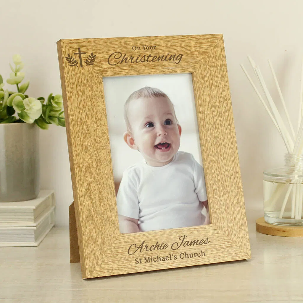 A wooden, landscape-oriented personalised photo frame for a Christening. The oak-effect wood is engraved at the top with a cross and foliage motif above the words "On Your Christening." Below the photo insert of a smiling baby (Archie James), the text is personalised with "Archie James" and "St Michael's Church." The frame is displayed on a light wooden surface next to a plant and a reed diffuser.