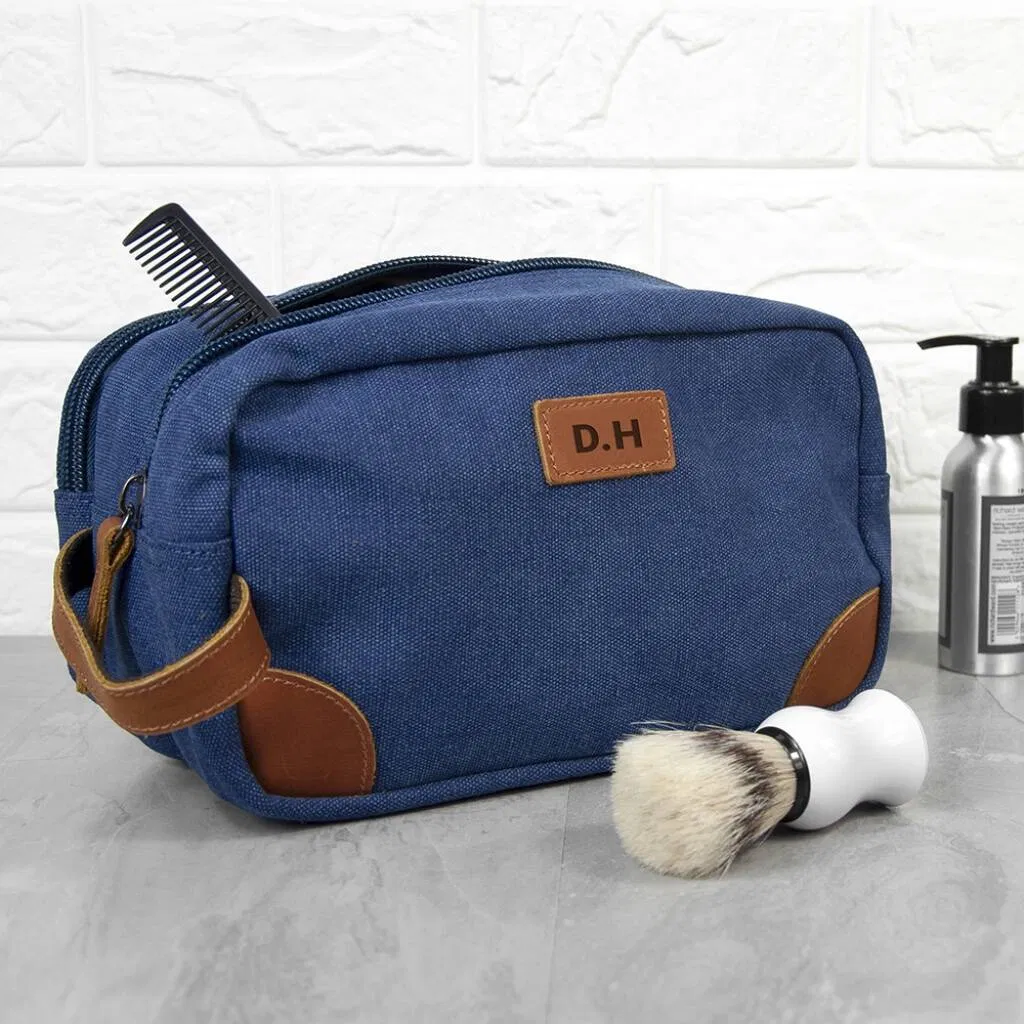 A blue denim wash bag with leather detailing and handles, featuring the initials 'D.H' on a leather patch. A black comb protrudes from the top of the bag, and a white shaving brush sits on the surface next to the bag.