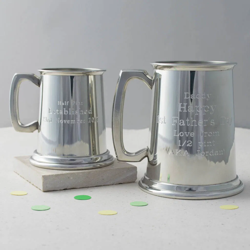 Two highly polished, silver-toned pewter tankards—one half-pint and one full-pint—engraved with personalised messages like 'Happy 1st Father's Day' and 'Established 21st November 2016'. The tankards are displayed on a white surface with confetti. Ideal traditional, engraved gift for Father's Day, Best Man, or Anniversary.