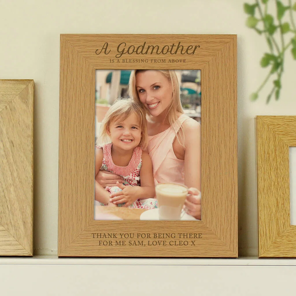Personalised wooden photo frame engraved with the fixed text "A Godmother is a blessing from above" at the top and custom personalised text below the photo, currently showing "Thank You for being there for me Sam, Love Cleo x". The frame holds a 5x7 portrait photo of a smiling woman holding a little girl.