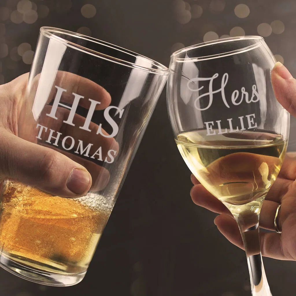 Close-up of a personalised pint glass engraved with HIS THOMAS toasting with a wine glass engraved with Hers ELLIE against a bokeh background.