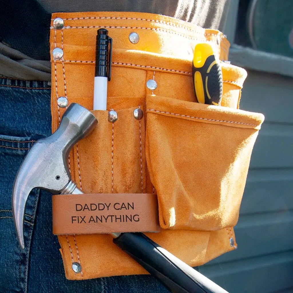 Personalised suede leather 11-pocket tool belt worn by a person in blue jeans, featuring a silver hammer, black marker pen, and a yellow screwdriver handle. A leather strap is embossed with the text: "DADDY CAN FIX ANYTHING."