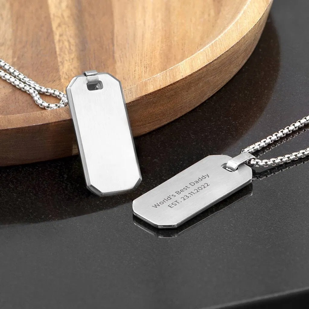 Close-up of two polished silver-tone men's dog tag necklaces, one engraved with the message 'World's Best Daddy EST. 23.11.2022', lying on a dark surface next to a wooden bowl.