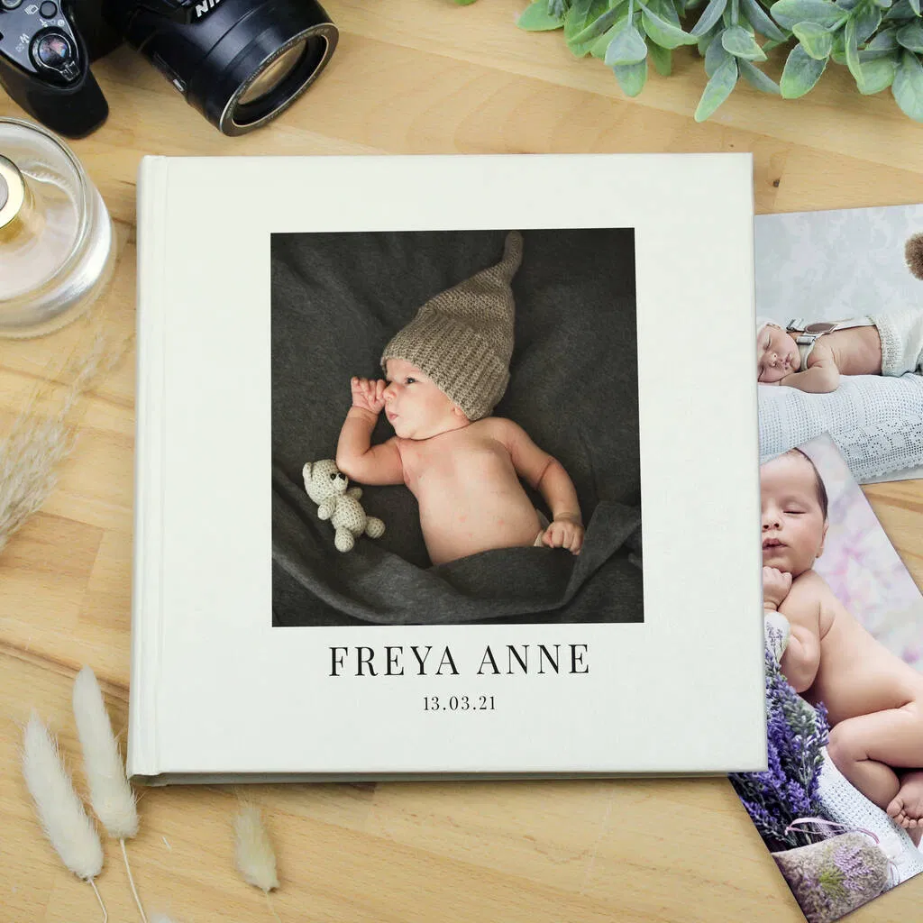 Overhead shot of a square, white, personalised new baby photo album with a clean, modern design resting on a wooden table. The cover features a main square photo of a newborn baby wearing a knit hat, with the personalised text "FREYA ANNE" and the date "13.03.21" printed below the photo. Other loose baby photos, a black camera, and a perfume bottle are visible nearby.