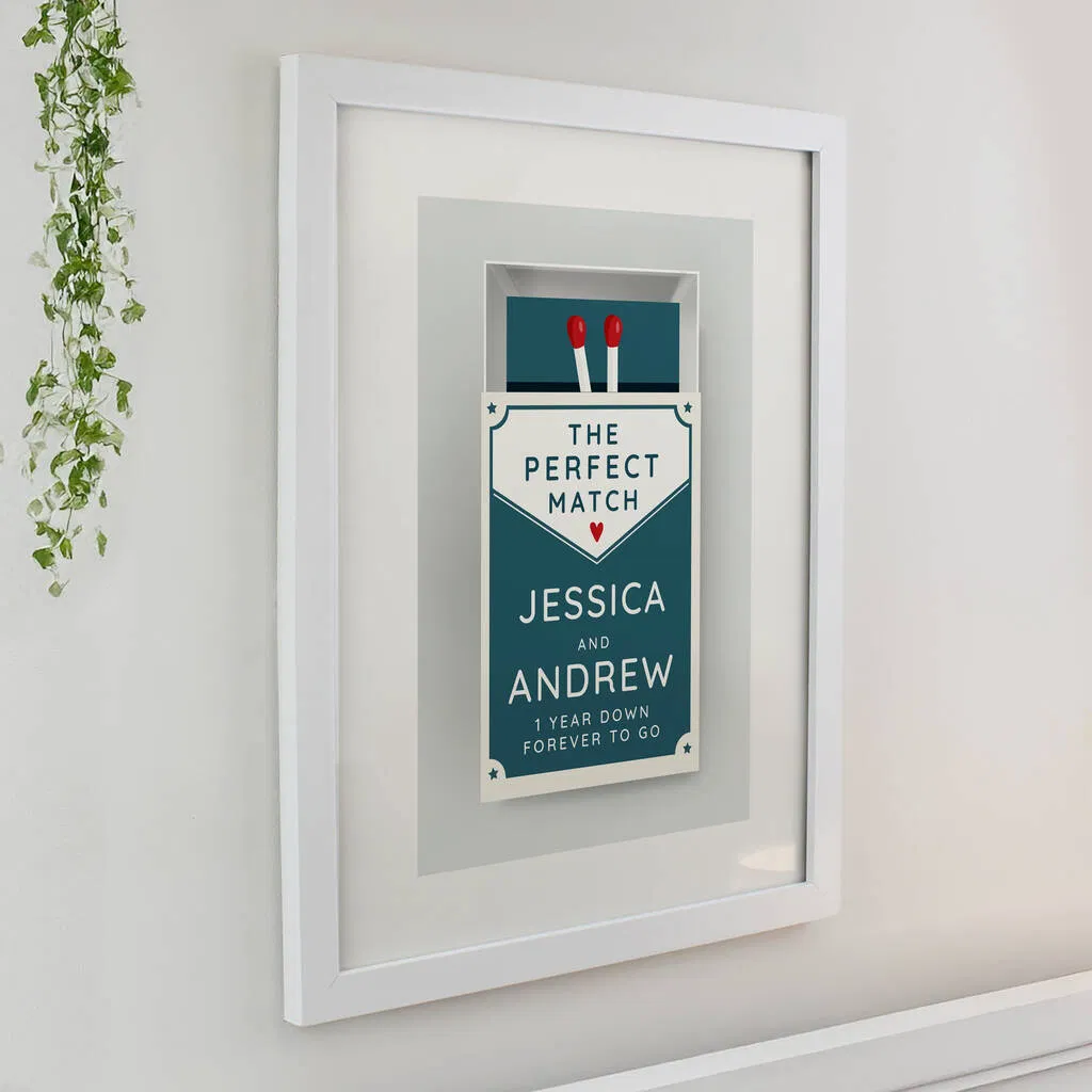 White framed wall art featuring a retro teal matchbox illustration with two red matches. Text reads THE PERFECT MATCH, followed by personalised names JESSICA AND ANDREW and a custom message 1 YEAR DOWN FOREVER TO GO.
