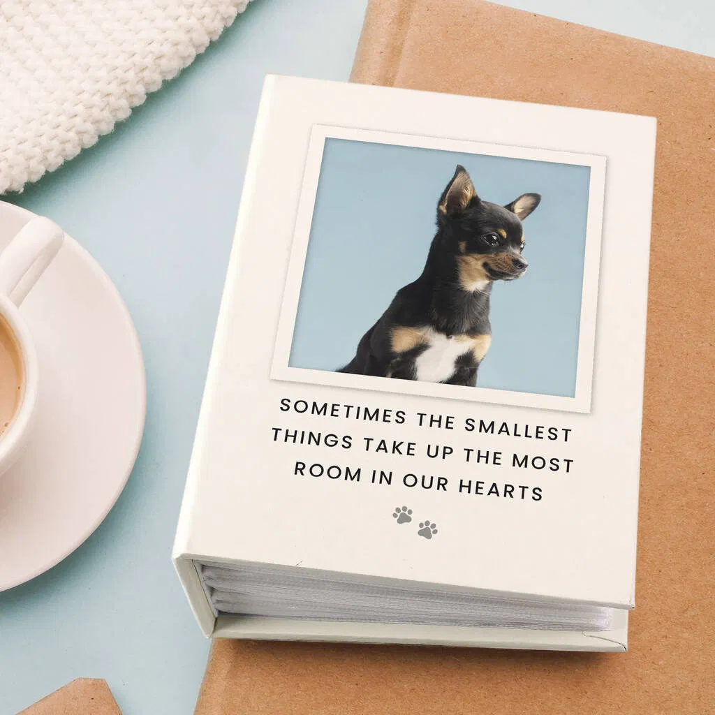 Overhead shot of a square, cream-colored personalised pet photo album with a custom cover. The cover displays a photo of a small black and tan Chihuahua and the text, "SOMETIMES THE SMALLEST THINGS TAKE UP THE MOST ROOM IN OUR HEARTS" with two paw print icons below the message. The album is resting on brown paper with a white coffee cup and knitting in the top left corner.