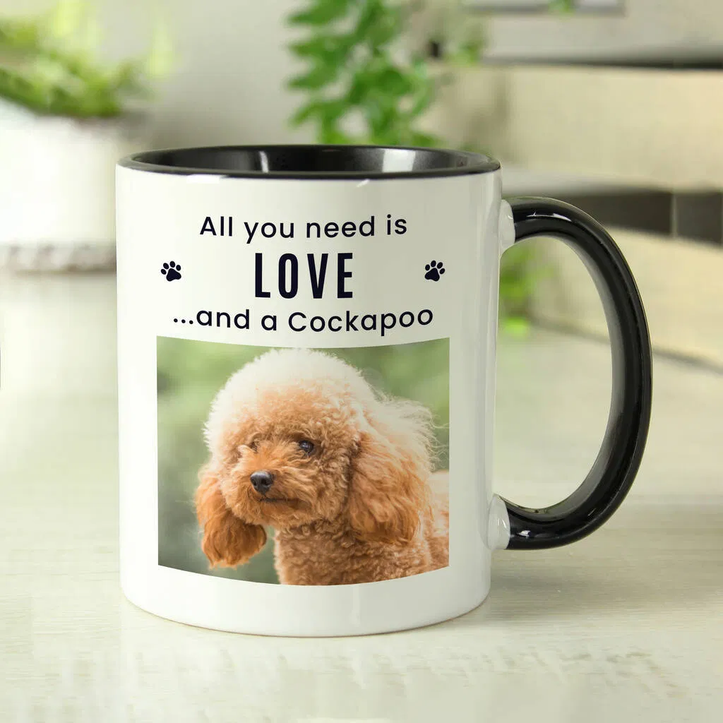 A white ceramic mug with a black handle and interior, personalised with a photo of a reddish-brown Cockapoo dog. Above the photo is the text, "All you need is LOVE...and a Cockapoo" flanked by paw print icons. The mug is sitting on a white wooden surface in a kitchen setting.