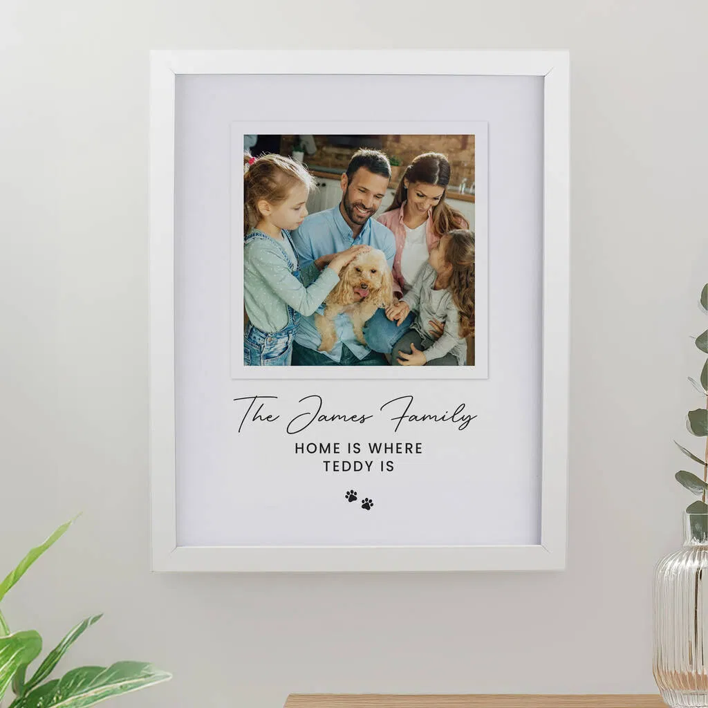 A white framed print displaying a high-quality photo of a happy family—a man, woman, and two young girls—cuddling a light brown Cocker Spaniel dog. Below the photo, the personalised text reads: "The James Family" in script font, and "HOME IS WHERE TEDDY IS" followed by two paw print icons. The frame hangs on a white wall above a wooden surface.