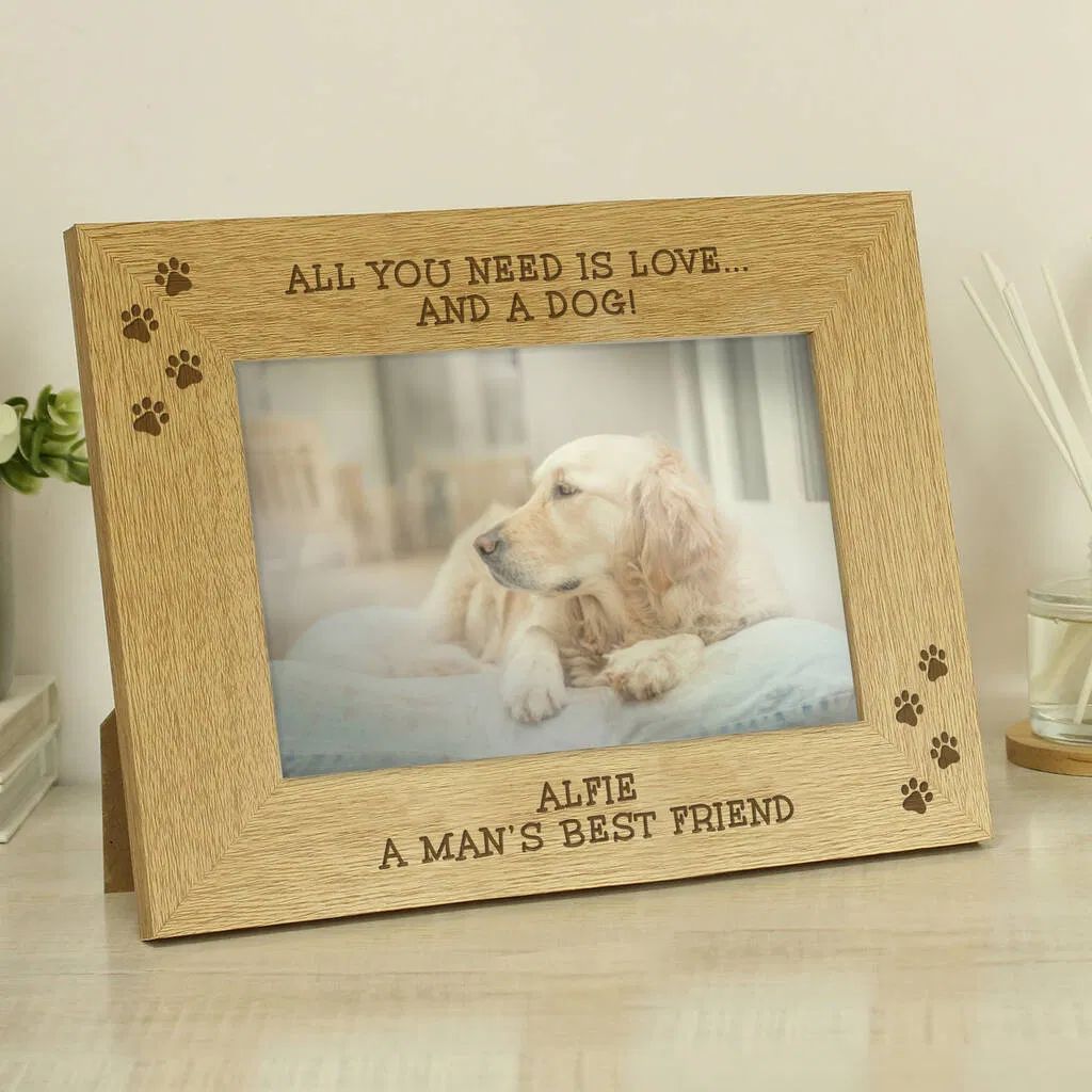A personalised wooden oak veneer picture frame designed for a landscape photo. The top is engraved with the text "ALL YOU NEED IS LOVE... AND A DOG!" flanked by paw print icons. The frame holds a 6x4 landscape photo of a light-coloured Golden Retriever lying down. Below the photo, the personalised text reads "ALFIE, A MAN'S BEST FRIEND" with more paw print icons.