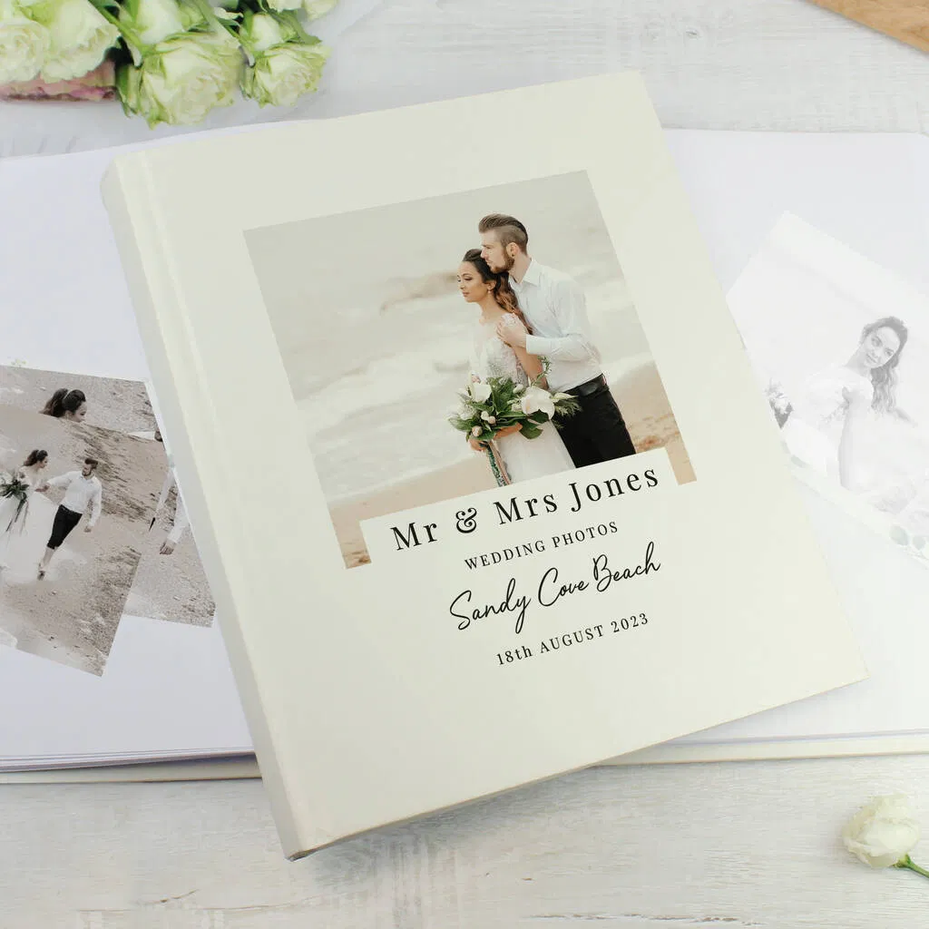 A square, cream-coloured personalised wedding photo album with a satin finish, featuring a photo of a married couple embracing on a beach. Below the photo, the cover is personalised with "Mr & Mrs Jones WEDDING PHOTOS Sandy Cove Beach 18th August 2023". The album is surrounded by white roses and other loose wedding photos.