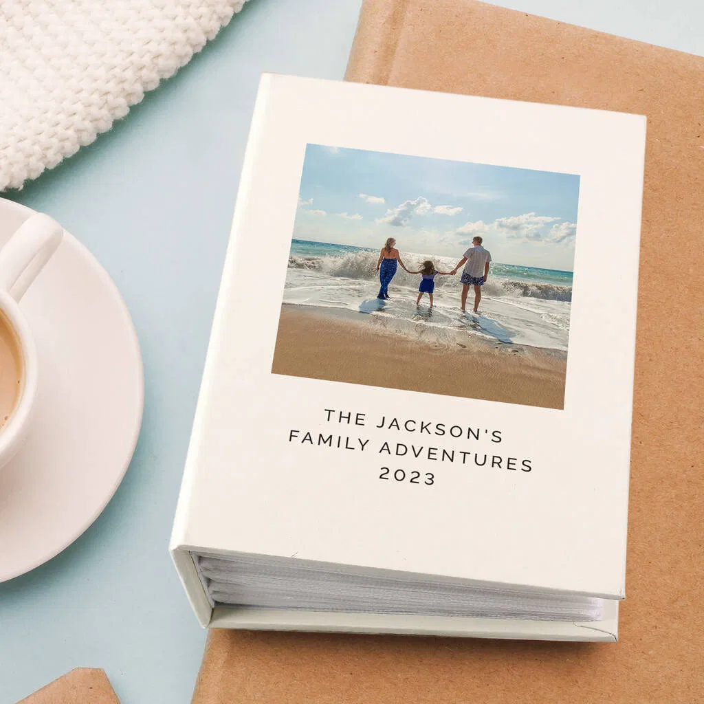 A square, cream-coloured personalised photo album with a custom cover featuring a photo of a family holding hands walking in the ocean surf on a sunny beach. Below the photo, the cover is personalised with the text "THE JACKSON'S FAMILY ADVENTURES 2023". The album is resting on brown paper with a white knitted blanket and a coffee cup in the corner.