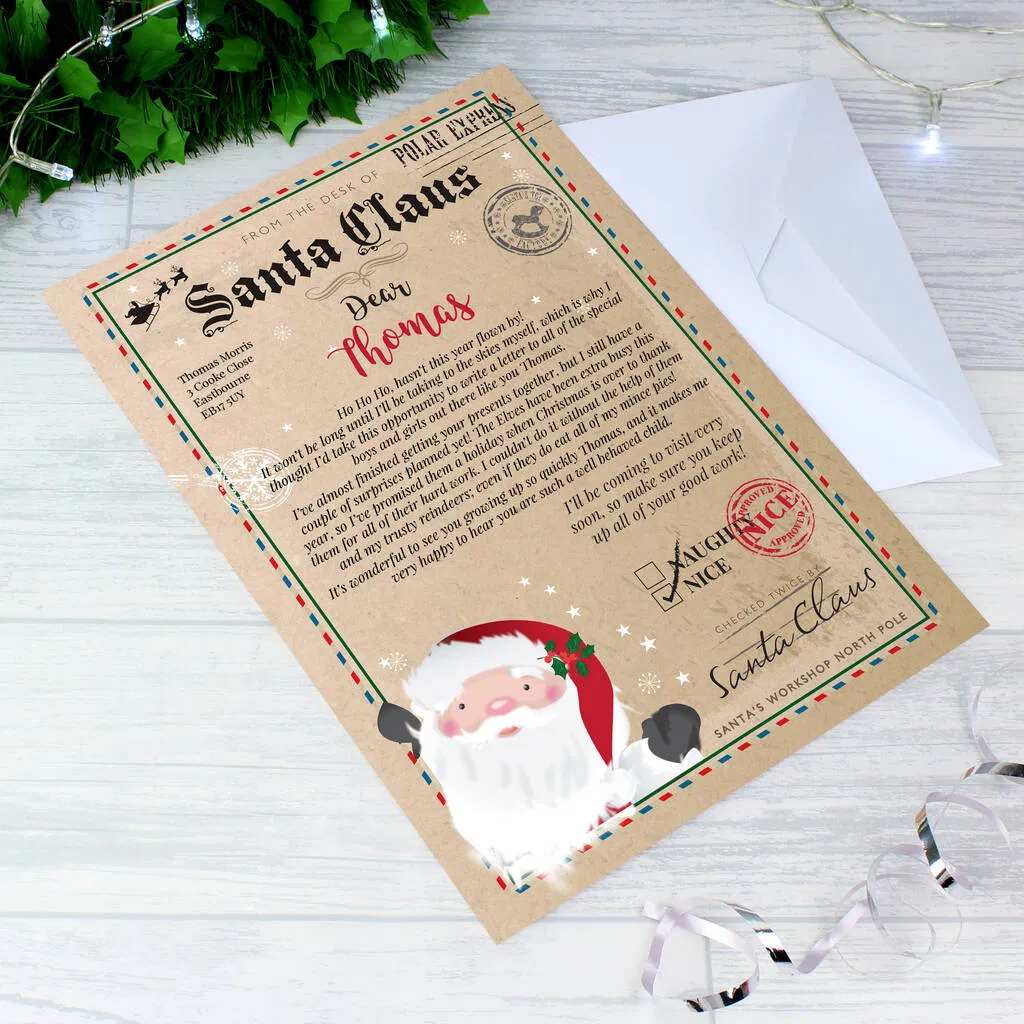 A personalised letter from Santa Claus on thick, vintage-style paper with festive borders, an image of Santa, and the recipient's name ("Thomas") clearly visible, laid diagonally on a wooden surface next to a white envelope and surrounded by subtle fairy lights and iridescent ribbon.