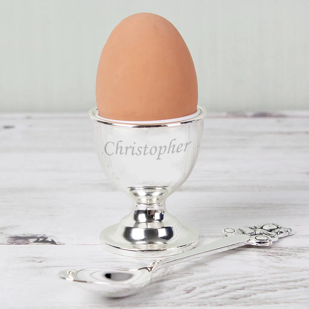 A personalised, polished silver-plated egg cup holding a whole brown egg, sitting on a pale wooden surface next to a matching silver spoon. The egg cup is engraved with the name 'Christopher' in a script font. Ideal traditional and keepsake gift for a child's Christening, first birthday, or Baptism.