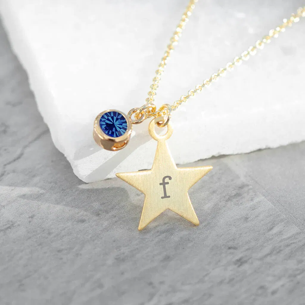 A gold-toned personalised necklace featuring a matte star charm engraved with a single lowercase letter ("f") and accompanied by a bezel-set, vibrant purple birthstone crystal charm, lying on a white marble surface next to a grey stone background.