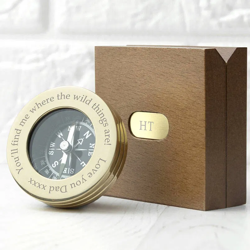 A personalised brass pocket compass resting next to its matching square, brown wooden presentation box, which features a monogrammed brass plate with the initials 'HT'. The compass bezel is engraved with the custom message 'You'll find me where the wild things are! Love You Dad xxxx'. This is an ideal luxury sentimental gift for a traveller, retirement, or graduation.