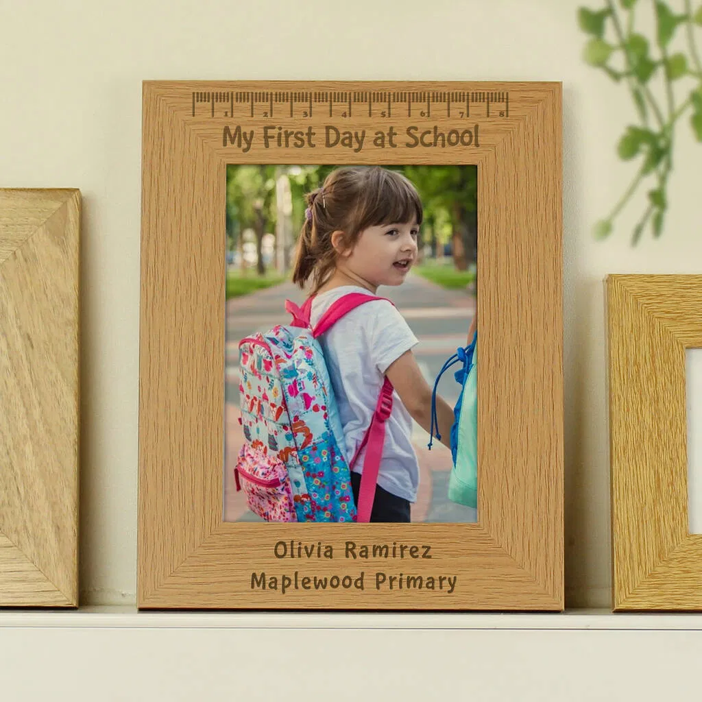 A light oak-coloured wooden photo frame holding a picture of a smiling young girl (like Olivia Ramirez) on her first day of school, wearing a colourful backpack. The frame is engraved with "My First Day at School" at the top, a ruler design, and a personalised name (e.g., Olivia Ramirez) and school name (e.g., Maplewood Primary) at the bottom.