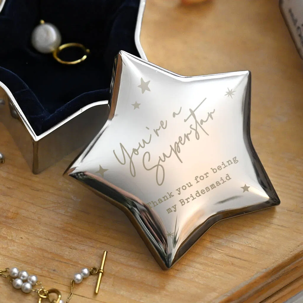 A nickel-plated, star-shaped trinket box with a reflective surface, engraved with the personalised message "You're a Superstar" and "Thank you for being my Bridesmaid," open to show a dark blue velvet interior holding a pearl ring and an earring.