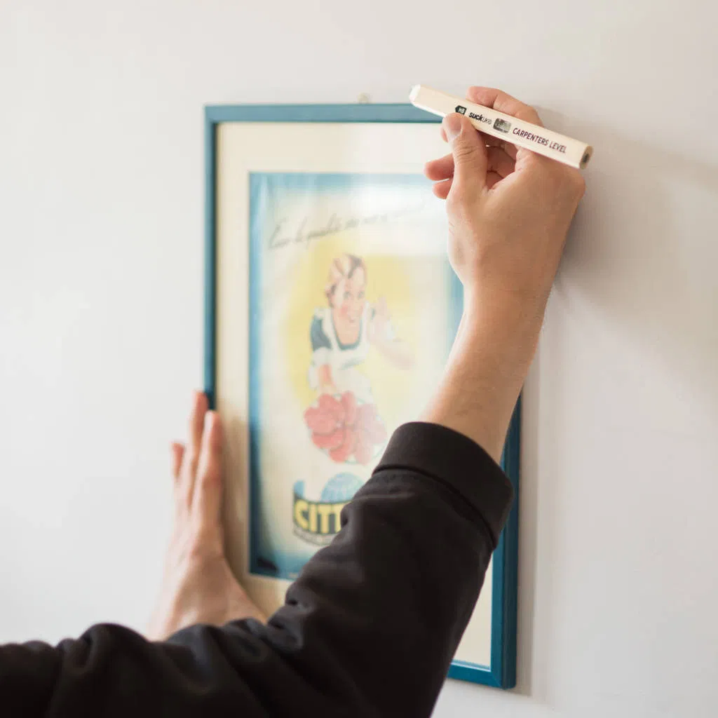 Hand using a novelty spirit level pencil to ensure a picture frame is hung straight on a white wall.