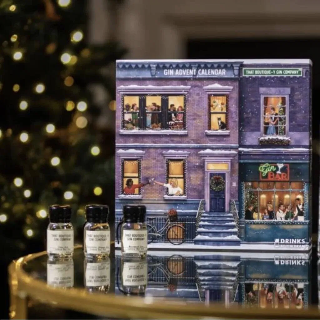 That Boutique-y Gin Company Advent Calendar 2025 displayed on a reflective surface in front of a subtly lit Christmas tree. The calendar is designed to look like a festive, snow-covered townhouse with windows showing various people celebrating, and a "Gin Bar" on the ground floor. Three miniature glass bottles of gin from the company are lined up in front of the calendar, reflecting on the surface.