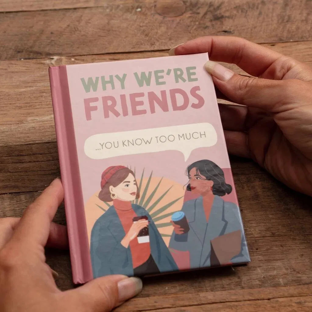 A small, pink and green fill-in-the-blank gift book titled 'Why We're Friends... You Know Too Much' being held open by a person's hands over a rustic wooden background. The cover features two illustrated women drinking coffee, emphasizing a close, humorous friendship. Ideal personalized gift for a best friend's birthday or Galentine's Day.